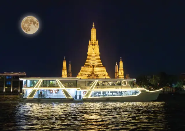 A lit-up boat passing Wat Arun under a full moon