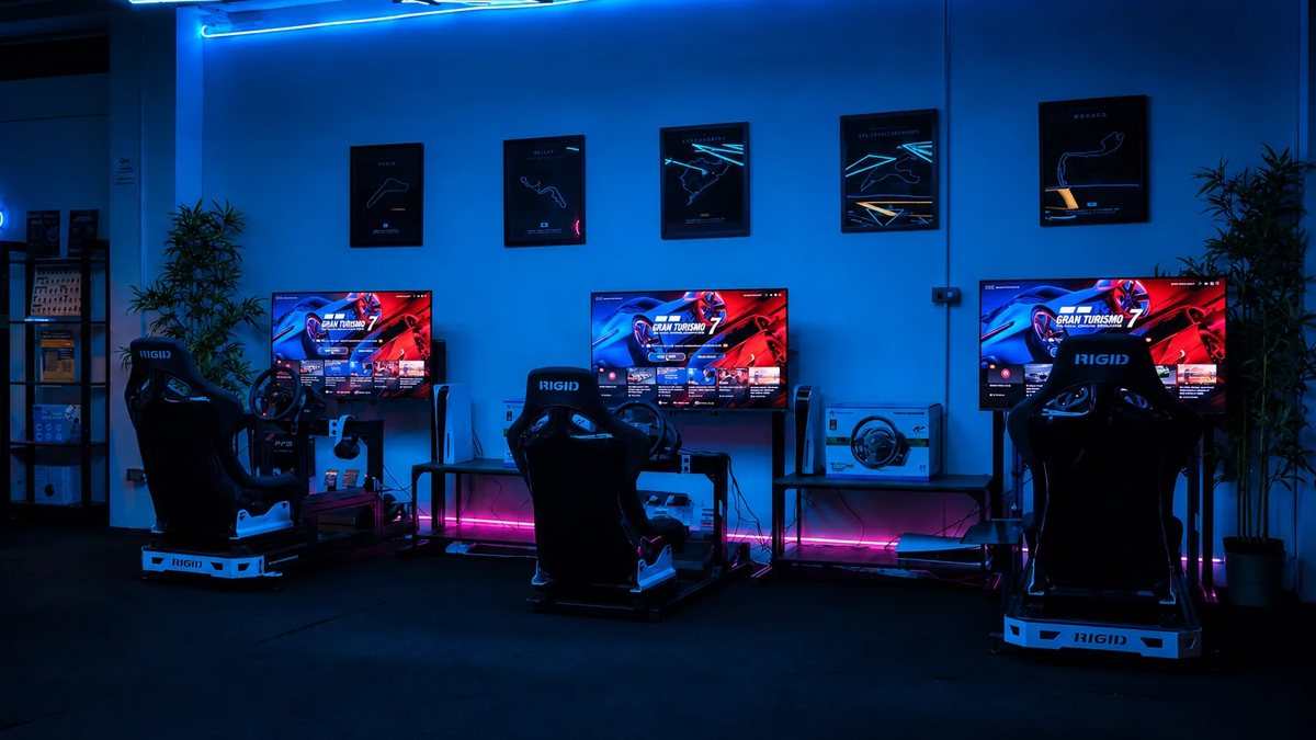 Three racing simulator seats with steering wheels and screens in a room lit blue