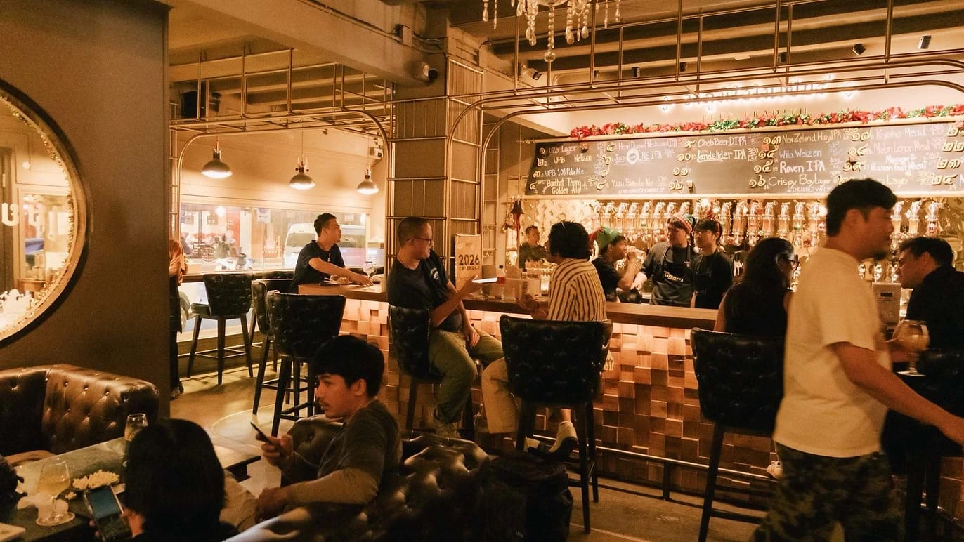 Drinkers at the bar and in leather armchairs at Beer Pradith, the tap list chalked on the wall behind