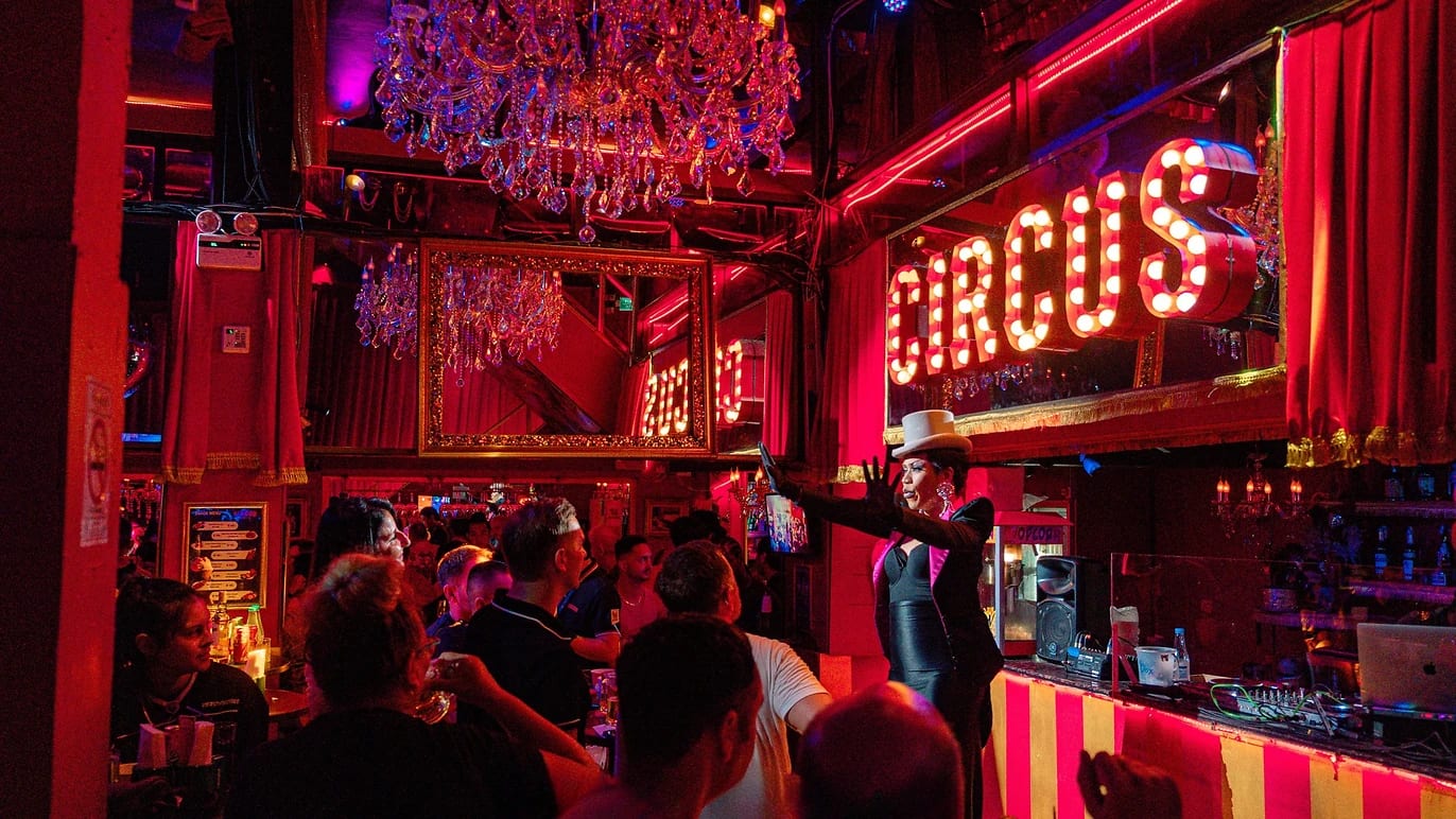 A drag performer in a top hat on stage under the Circus sign and a chandelier, the crowd watching