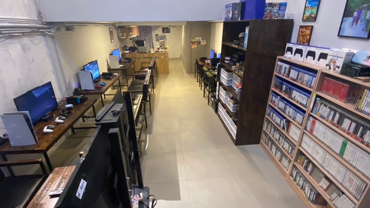 Consoles on desks along one wall of Game Bar Odyssey, facing shelves full of game boxes