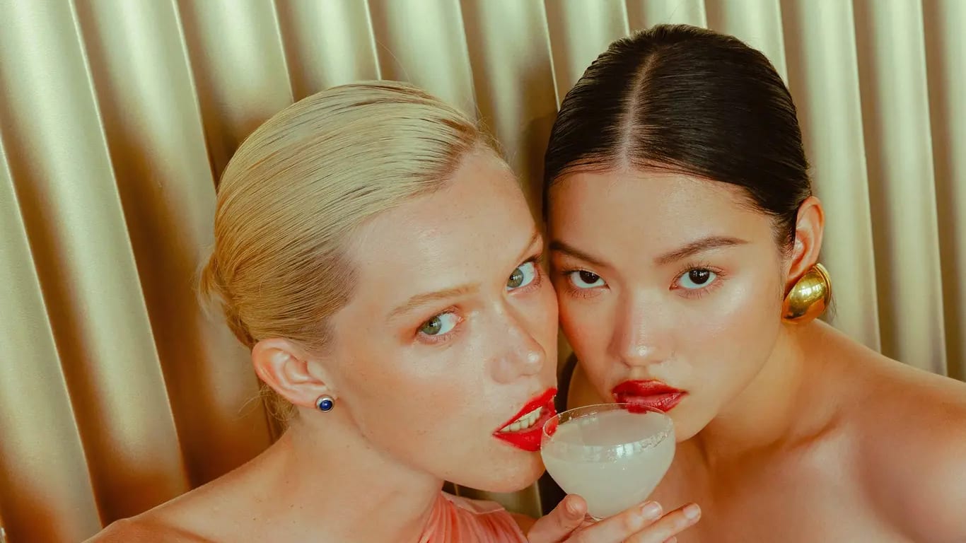 Two women sharing one cocktail in front of a gold curtain, from The Standard's bar crawl campaign