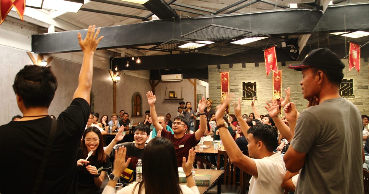 A packed game night at The Stronghold board game café in Siam Square, players raising their hands